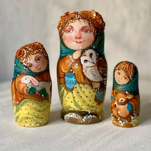 Pode incluir: Um conjunto de três bonecas russas de madeira pintadas à mão. A boneca maior está vestindo um casaco marrom, uma echarpe azul e um vestido amarelo com bolinhas brancas. Ela está segurando uma coruja branca. A boneca do meio está vestindo um casaco marrom, uma echarpe verde e um vestido amarelo com bolinhas brancas. Ela está segurando um coelho branco. A boneca menor está vestindo um casaco marrom, uma echarpe azul e um vestido amarelo com bolinhas brancas. Ela está segurando um veado marrom.