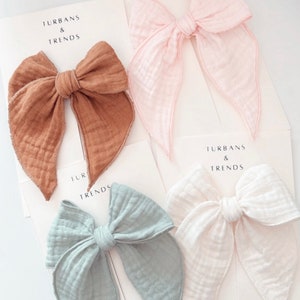 Puede incluir: Cuatro lazos para el pelo de tela en diferentes colores. Los lazos están hechos de una tela suave y texturizada y están atados en un nudo grande y suelto. Los lazos están dispuestos sobre una superficie blanca con el texto "Turban & Trends" impreso en cada tarjeta.