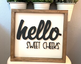 May include: A white wooden sign with a black cursive "hello sweet cheeks" message. The sign is framed with a brown wood frame and is sitting on top of a white toilet.