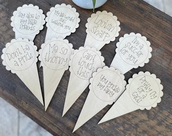 May include: Eight wooden plant markers with scalloped edges and humorous sayings. The sayings include "Say aloe to my little friends", "My life would succ without you", "Don't be a prick", "Me so thorny", "Lookin' sharp", "Can't touch this", "I'm a succ-er for you", "You prickle my lancy", and "Pretty fly for a cacti".