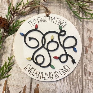May include: A round wooden ornament with a black string for hanging. The ornament features a black silhouette of tangled Christmas lights with colored bulbs. The text "It's fine, I'm fine, everything is fine!" is written around the lights.