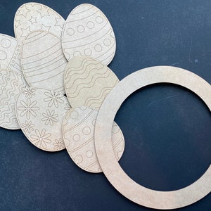 May include: A selection of unfinished wooden Easter egg cutouts with etched patterns, such as stars, dots, and flowers. A wooden circular frame is also present. The items are displayed on a dark surface.