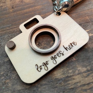 May include: A wooden camera keychain with a cutout for a logo. The keychain is light brown with a dark brown wood accent. The text "logo goes here" is engraved on the keychain.