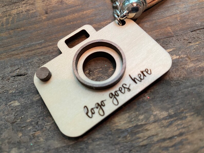 May include: A wooden camera keychain with a cutout for a logo. The keychain is light brown with a dark brown wood accent. The text "logo goes here" is engraved on the keychain.