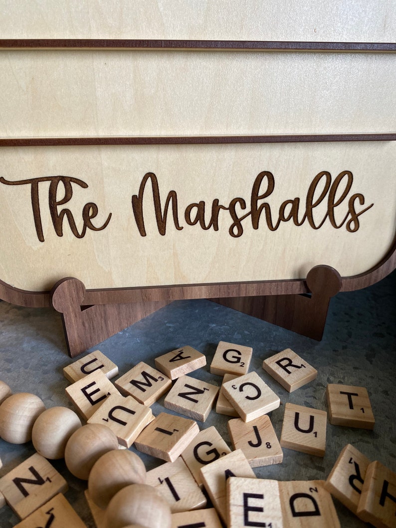 May include: A wooden sign with the text "The Marshalls" engraved on it. The sign is sitting on a wooden stand and there are wooden letter tiles scattered around it.