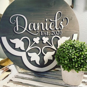 May include: A round wooden sign with the name "Daniel's" written in white cursive lettering. The sign also includes the text "Est. 2020" and a white floral design.