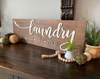 May include: A rectangular wooden sign with the word "laundry" in white cursive lettering. Below, the words "wash, dry, fold" are printed. The sign is brown and is decorated with decorative items.