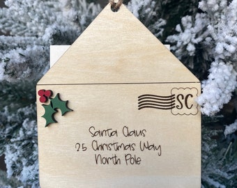 May include: A wooden Christmas ornament shaped like an envelope with a holly sprig and red berries. The address reads "Santa Claus, 25 Christmas Way, North Pole".