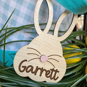 Puede incluir: Etiqueta de cesta de Pascua de madera en forma de conejo con el nombre Garrett escrito en cursiva. La etiqueta es de color marr&oacute;n claro con una nariz rosa y bigotes negros.