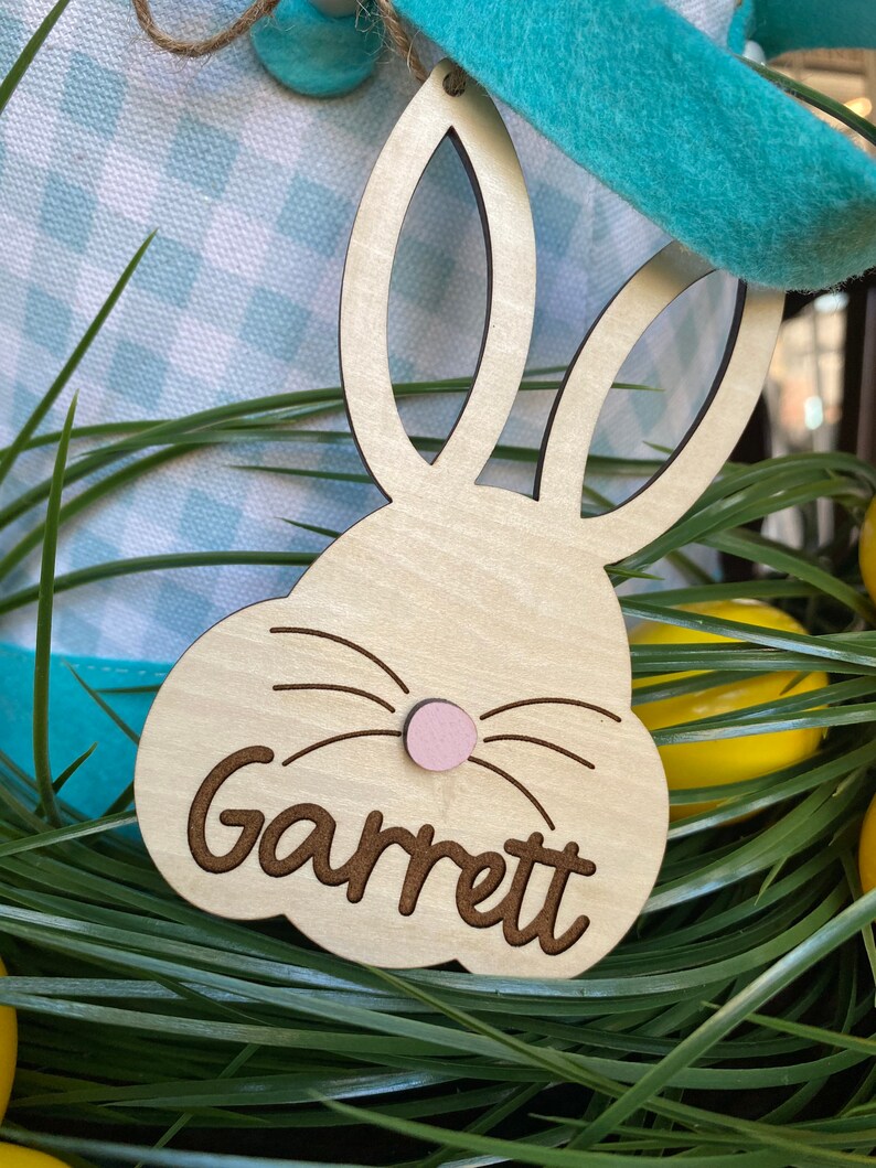 Puede incluir: Etiqueta de cesta de Pascua de madera en forma de conejo con el nombre Garrett escrito en cursiva. La etiqueta es de color marr&oacute;n claro con una nariz rosa y bigotes negros.
