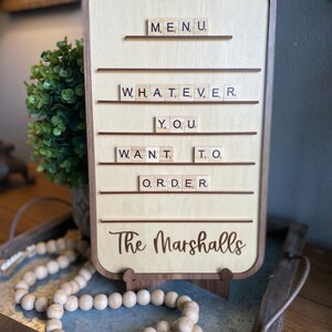 May include: A wooden sign with a brown frame and a light wood background. The sign has a scrabble tile message that reads "MENU, WHATEVER YOU WANT TO ORDER." The sign also has the text "The Marshalls" in brown cursive lettering.