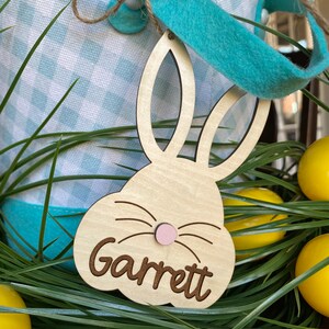 Puede incluir: Una etiqueta de cesta de Pascua de madera con forma de conejo con el nombre "Garrett" grabado en ella. La etiqueta est&aacute; unida a una cesta de Pascua a cuadros azules y blancos con una cinta azul.