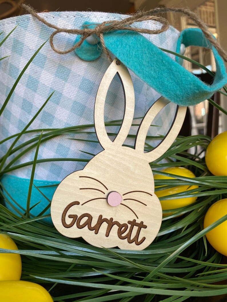 Puede incluir: Una etiqueta de cesta de Pascua de madera con forma de conejo con el nombre "Garrett" grabado en ella. La etiqueta est&aacute; unida a una cesta de Pascua a cuadros azules y blancos con una cinta azul.
