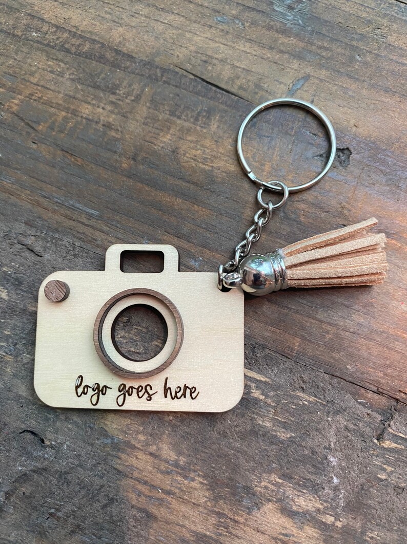 May include: A wooden camera keychain with a brown tassel. The keychain has a space for a logo with the text "logo goes here" engraved on the bottom.