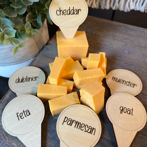May include: Wooden cheese markers with different cheese names written on them. The names include cheddar, dubliner, feta, parmesan, muenster, and goat. The markers are placed around a variety of cheese cubes.