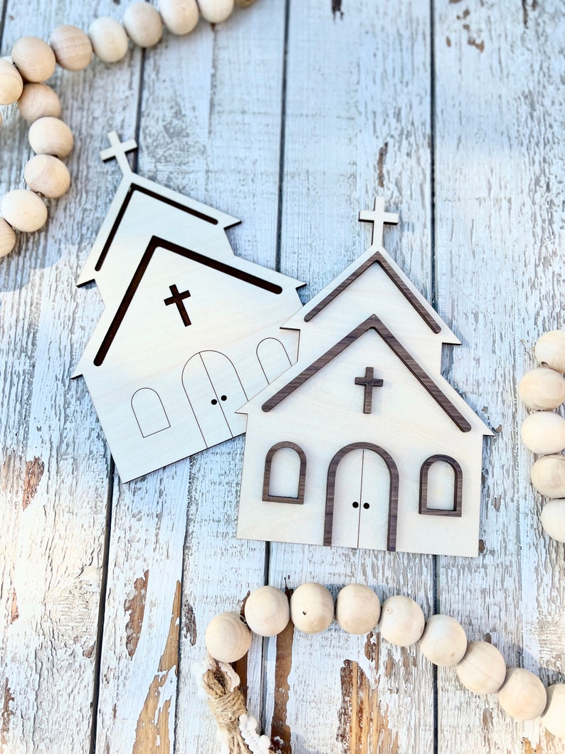 May include: Two wooden church cutouts with a brown stain on a whitewashed wood background. The church cutouts have a cross on the top and windows and a door.
