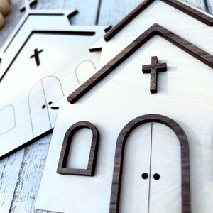 May include: Two wooden church cutouts with brown trim. The larger cutout has a cross on the front and two arched doors with black door knobs. The smaller cutout has a cross on the front and two arched windows.
