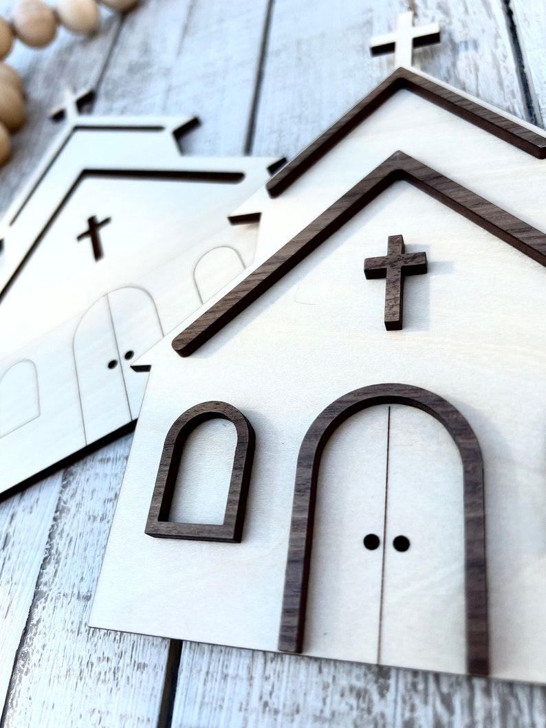 May include: Two wooden church cutouts with brown trim. The larger cutout has a cross on the front and two arched doors with black door knobs. The smaller cutout has a cross on the front and two arched windows.