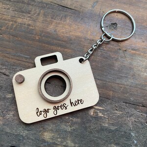 May include: A wooden camera keychain with a metal ring and chain. The camera has a cutout for a logo and the text "logo goes here" is engraved on the front.