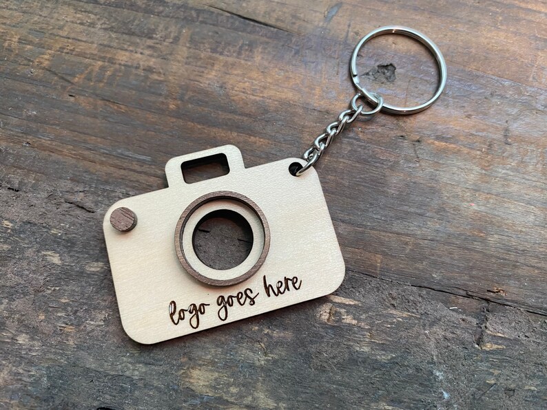 May include: A wooden camera keychain with a metal ring and chain. The camera has a cutout for a logo and the text "logo goes here" is engraved on the front.