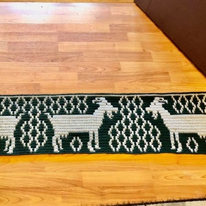 May include: A green and white crocheted rug with a repeating pattern of goats. The rug has fringe on both ends.