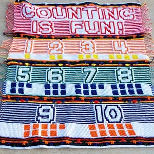 Peut inclure: Textile fait à la main et coloré avec l'inscription "COUNTING IS FUN!" et les chiffres de 1 à 10. Chaque chiffre est affiché sur une bande séparée avec une palette de couleurs unique, comprenant le rouge, l'orange, le vert, le bleu et le blanc. Les bords sont frangés.