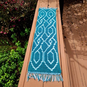 Puede incluir: Una bufanda de ganchillo turquesa y blanca con un patrón geométrico. La bufanda es rectangular con un diseño blanco repetido sobre un fondo turquesa. Los extremos tienen flecos blancos y turquesas.