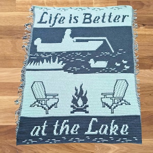 Könnte beinhalten: Eine gehäkelte Decke in Blau- und Grüntönen. Die Decke zeigt den Text "Life is Better at the Lake" und Bilder von einem Boot, einem Lagerfeuer und zwei Stühlen.