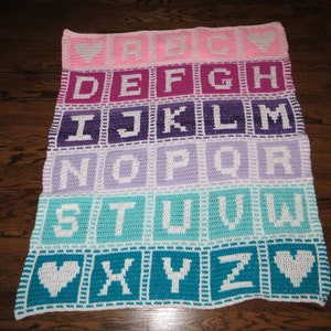 May include: A crocheted alphabet blanket with a rainbow color gradient. The blanket features white letters in each square, with hearts at the beginning and end of the alphabet.