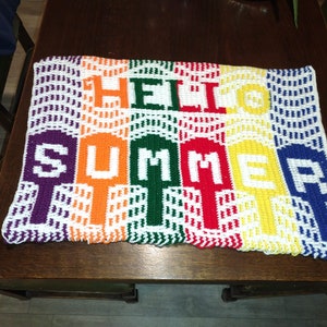 May include: A vibrant, crocheted banner displaying "HELLO SUMMER" in rainbow colours. The bold, block letters are set against a white background with a wavy border. The banner is placed on a dark wooden table.