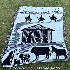 May include: A black and white woven blanket featuring a nativity scene. The blanket depicts the three wise men on camels, a stable with Mary, Joseph, and baby Jesus, and various animals. The text "Crocheted by Sue Lemal (front)" is at the bottom.
