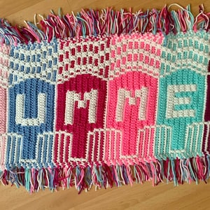 May include: A vibrant, hand-crocheted rug spelling "SUMMER" in white letters. The design features popsicle shapes in pink, blue, and teal, with a fringed border. The rug is a decorative item.