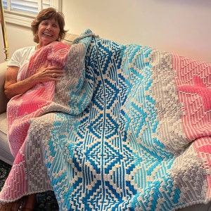 May include: A woman sits on a couch with a large, colorful crocheted blanket. The blanket features a geometric pattern in shades of pink, blue, and gray.