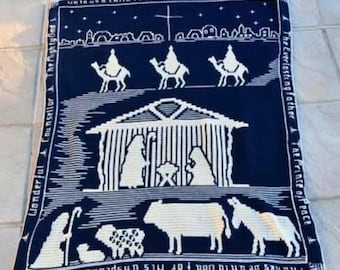 May include: A navy blue and white crocheted blanket featuring a nativity scene. The design includes the holy family, wise men, animals, and text. The blanket has a border with additional text. The text includes "Wonderful Counselor" and "Thanks be unto God for His unspeakable gift."