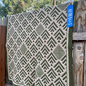 May include: A green and beige crocheted blanket with a geometric pattern of triangles. A blue ribbon with the text "First Place, Colorado State Fair, Pueblo, CO" is attached to the blanket.