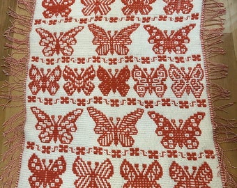 May include: A white crocheted throw blanket with a fringe border features a repeating pattern of orange butterflies. The blanket is made with a simple stitch and has a rustic, handmade look.