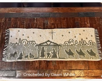 May include: A rectangular, off-white crocheted tapestry with a nativity scene design in dark green. The tapestry features a cross, stars, and silhouettes of camels, figures, and buildings. The text "Crocheted by Dawn White" is at the bottom.