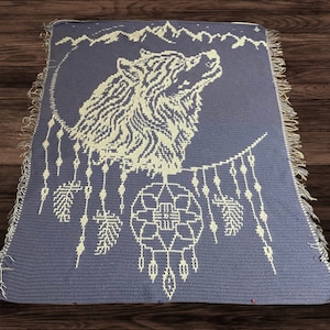 Könnte beinhalten: Eine gehäkelte Wolldecke mit einem weißen Wolf, der zum Mond heult, und einem Traumfänger-Design. Die Decke ist helllila mit weißen Akzenten.