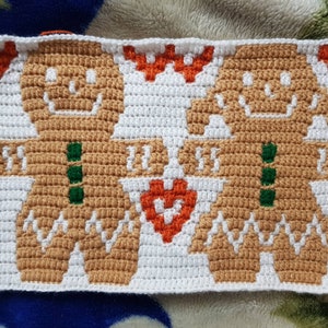 Puede incluir: Diseño navideño tejido a crochet con dos hombrecitos de jengibre sosteniendo un corazón. Las figuras de jengibre son de color marrón claro con botones verdes y detalles blancos, sobre un fondo blanco con elementos decorativos naranjas.