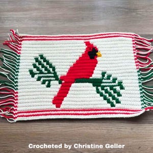 Puede incluir: Una pieza rectangular tejida a ganchillo con un cardenal rojo sobre una rama verde sobre un fondo blanco. Tiene flecos rojos y verdes y un borde rojo. El texto "Crocheted by Christine Geller" está en la parte inferior.