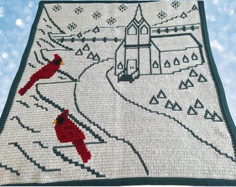 May include: A crocheted blanket featuring a winter scene with a church, road, and two red cardinals. The blanket has a cream background with dark green detailing and border. The text "Crocheted by Pamela Beckford" is at the bottom.