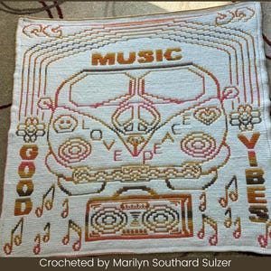 May include: A crocheted blanket with a white background and colorful designs. The blanket features a peace sign, a van, a cassette tape, musical notes, and the words "Good Vibes", "Love", "Peace", and "Music".