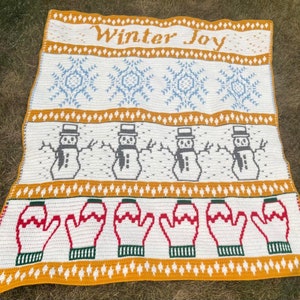 May include: A white crocheted blanket with a winter theme. The blanket features a repeating pattern of snowflakes, snowmen, and mittens. The words "Winter Joy" are written in yellow yarn at the top of the blanket.