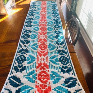 Puede incluir: Un camino de mesa tejido a crochet con un patrón geométrico en tonos de azul, turquesa, blanco y coral. El camino de mesa está colocado sobre una mesa de madera.