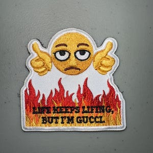 May include: Embroidered patch featuring a yellow emoji with a sad expression and thumbs up. The emoji is surrounded by flames and the text "LIFE KEEPS LIFTING, BUT I'M GUCCI" in black letters.