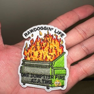 May include: Embroidered patch featuring a green dumpster engulfed in flames. The patch reads "RAWDOGGIN' LIFE" above the dumpster and "ONE DUMPSTER FIRE AT A TIME" below. The flames are orange and yellow.