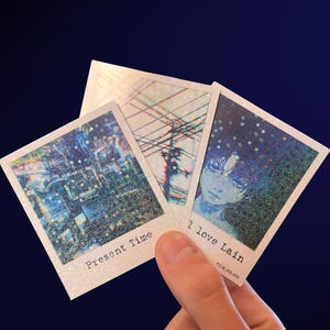 May include: Three holographic art prints are held against a dark blue background. The prints feature images of a cityscape, power lines, and a portrait. Text on the prints reads "Present Time" and "I love Lain".