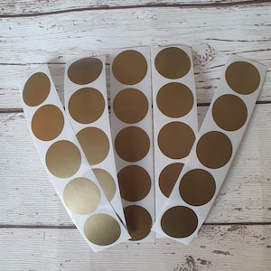 May include: Five vertical strips of adhesive labels, each featuring a series of circular gold stickers. The stickers have a smooth, metallic finish and are arranged in a single column on a white backing paper, creating a set of uniform round labels.