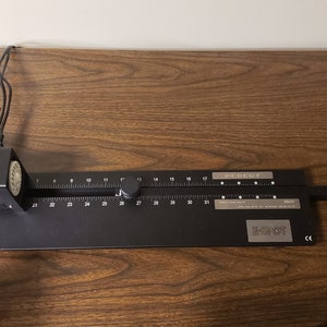 May include: A black and silver DECUT cutting machine with a ruler that goes up to 31 inches. The machine is on a wooden surface. The machine has a black arm that extends to the right. The machine has a white and black control panel on the left side.