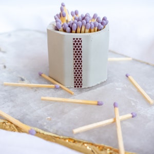 Puede incluir: Un porta-cerillas de hormigón gris con un patrón de panal en la parte delantera. El porta-cerillas está lleno de cerillas con puntas moradas. El porta-cerillas está sobre una superficie blanca.
